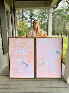 a woman standing behind two pink paintings on a porch with trees and birds painted on them