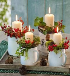 three white mugs filled with red berries and greenery sit on a tray next to candles