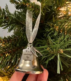 a hand holding a silver bell ornament in front of a christmas tree with writing on it