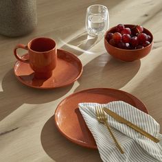 two orange plates with silverware and fruit on them sitting on a table next to a bowl of cherries