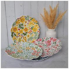 three plates with flowers on them next to a white vase and dried grass in the background