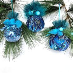 three blue ornaments hanging from a pine branch with green leaves and balls on them, all decorated in shades of blue