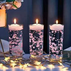 three lit candles with pink flowers in them on a table next to a flower arrangement