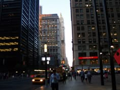 people are walking on the sidewalk in front of tall buildings at dusk, with one car driving down the street