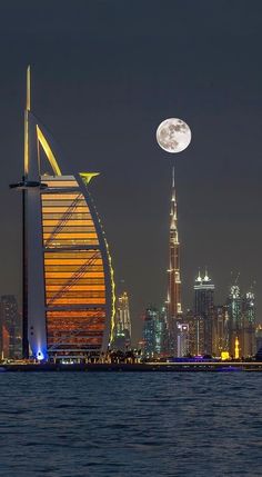 the full moon is setting in front of a tall building with a sailboat on it's side