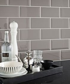 a kitchen counter with dishes, cups and bottles on it in front of a gray tiled wall