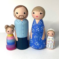 three wooden dolls are standing next to each other on a white surface, one is wearing a blue dress