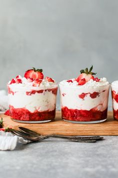 three desserts with strawberries and whipped cream in them on a wooden board next to silverware