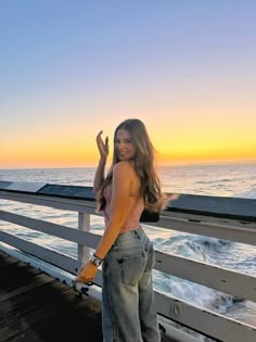 a beautiful young woman standing on top of a pier next to the ocean at sunset