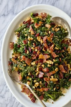 a white bowl filled with greens and bacon on top of a marble countertop next to two spoons