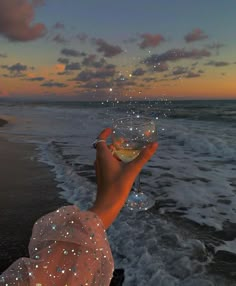 a person holding a wine glass in front of the ocean at sunset with bubbles coming out of it