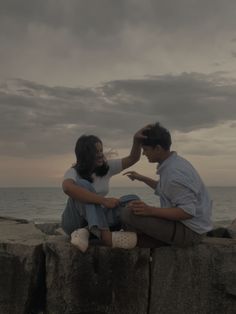 a man and woman sitting on rocks near the ocean with their hands touching each other's foreheads