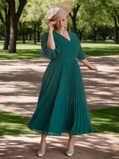 a woman in a green dress and hat poses for the camera with trees in the background