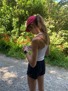 a woman in shorts and a red hat is looking at her cell phone while standing on a gravel road