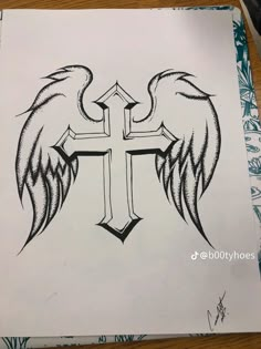 a cross with wings drawn on paper next to a pen and ink drawing pencils