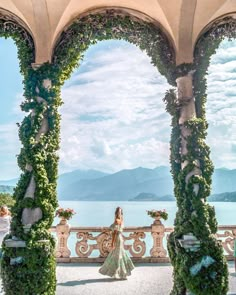 a woman in a white dress standing under an arch covered with greenery near the water