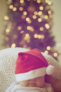 a baby wearing a santa hat sitting in front of a christmas tree with lights on it