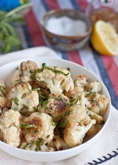 a white bowl filled with cauliflower sitting on top of a table next to lemon wedges