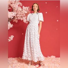 a woman standing in front of a pink background wearing a white dress with flowers on it