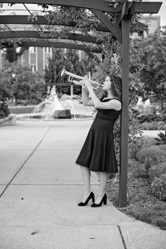 a woman in black dress playing trumpet under a pergolated arbor with fountain behind her