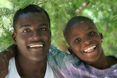 two men are smiling for the camera in front of some trees and bushes, one is holding his arm around the other man's shoulder