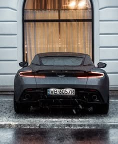 the rear end of a grey sports car parked in front of a window with curtains