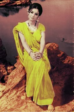 a woman sitting on top of a rock next to a body of water in a yellow dress