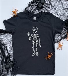 "Spooky, scary skeletons Send shivers down your spine.... Spooky scary skeletons Give you the peace sign! Our remix of that song is now in t-shirt form! This fun skater skeleton tee is destined to be your kid's new halloween favorite!  Bad 2 the Bone is made using a 100% cotton t-shirt in the color \"vintage black\" WASH & CARE INSTRUCTIONS: To maintain the integrity of the shirt, we suggest washing inside out in cold water with like colors, using a mild/color safe detergent. Dry on low heat or Black Horror Tops With Skull Print, Unisex Black Tops With Skull Print, Funny Halloween Top With Skull Print, Funny Halloween Tops With Skull Print, Funny Halloween Skull Print Top, Funny Skull Print Tops For Halloween, Halloween Fun Skull Print T-shirt, Unisex Halloween T-shirt, Halloween Glow In The Dark Black Top