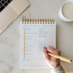 a person is holding a pencil and writing on a planner next to a cup of coffee