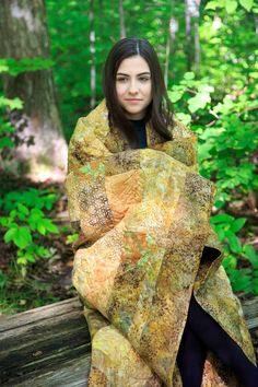 a woman is sitting on a log in the woods with a blanket over her head