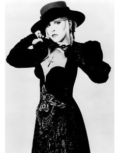 a woman in a black dress and hat posing for the camera with her hands on her head