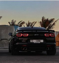 the rear end of a black sports car parked in front of a concrete wall and palm trees