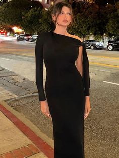 a woman in a black dress standing on the side of a road with her hand on her hip