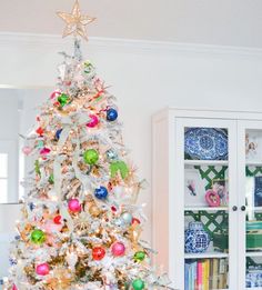 a white christmas tree with colorful ornaments in the corner and a china cabinet behind it