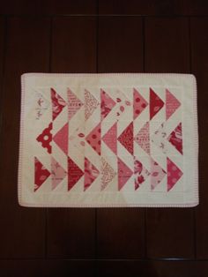 a quilted table runner with red and white triangles on it's side, sitting on a wooden floor