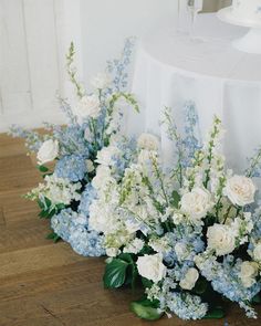 some white and blue flowers are on the floor next to a round cake stand with a table cloth