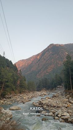 a river running through a forest filled with rocks and power lines in the mountains behind it