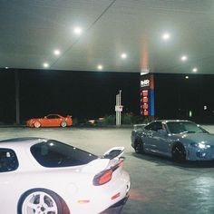 two sports cars parked in a parking lot at night with gas pumps and lights above them