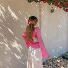 a woman standing in front of a white wall wearing a pink cardigan and long white skirt