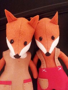 two stuffed foxes are sitting next to each other on a shelf together, one is wearing a red dress and the other has a pink skirt