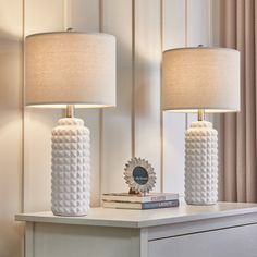 two white lamps sitting on top of a table next to a book and lamp shade