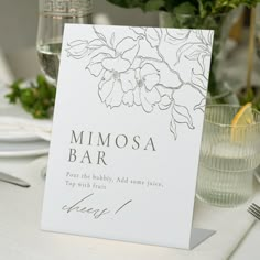 a place card with flowers on it sitting on a table next to glasses and silverware