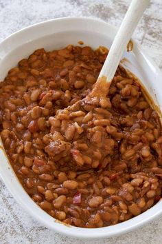 a white bowl filled with baked beans sitting on top of a counter next to a wooden spoon