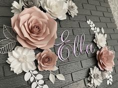 the name ella is surrounded by paper flowers and leaves on a gray brick wall with silver accents