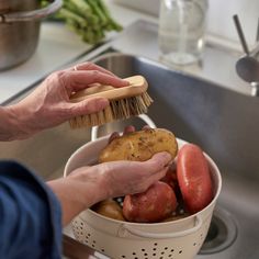 a person holding a brush over a bucket filled with potatoes and other vegetables in a kitchen sink