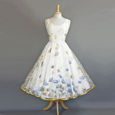 A striking tea length wedding dress made from a charming, embroidered lace featuring blue poppies and delicate white and gold daises. It features a classic darted scoop neck bodice and a flattering fit and flare silhouette with a tea length circle skirt edged with gold satin trim. The dress is made from a layer of lace over an Ivory Cotton fabric which is then lined.  Our darted bodice is not boned at all, and so it needs to be worn with a supportive bra to give you the most flattering fit. PRODUCTION TIMES & SHIPPING.  This dress is made to order in the UK by one of our sewers. It can be made in one of our standard sizes or made-to-measure so please bear in mind that the dress will take approximately 6-12 weeks to make and 1-3 weeks for international shipping, depending on location. For a 1950s Tea Length Wedding Dress, 1950 Wedding Dress, 1950s Wedding Dress Tea Length, 1950 Wedding, Dig For Victory, Blue Poppies, Dress Tea Length, Lace Bolero, Tea Length Wedding