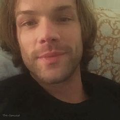 a man with long hair sitting on a couch looking at the camera while wearing a black shirt