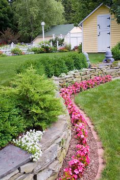 a garden with flowers and rocks in the grass near a yellow house that has a white picket fence around it