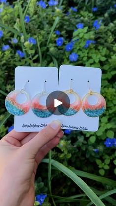 the earrings are being held up in front of some blue and green flowers, with one person's hand holding it