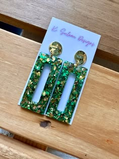 two green and gold earrings sitting on top of a wooden bench